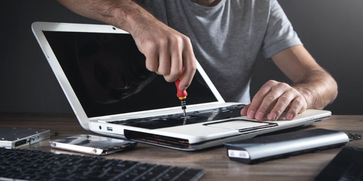 A technician uses a screwdriver to repair a disassembled laptop on a desk, with tools and parts spread around