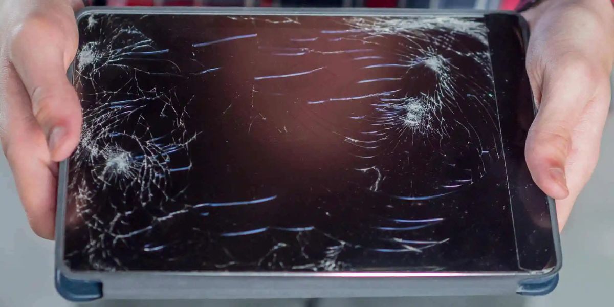 Hands holding a tablet with multiple cracks and spiderweb patterns across the damaged screen