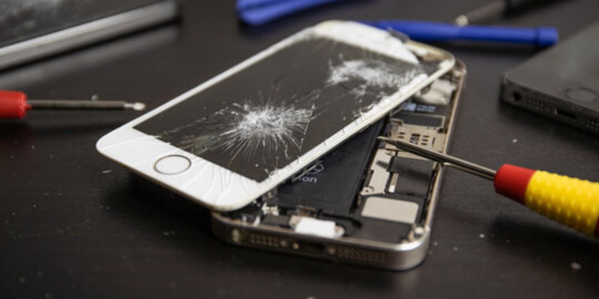 Cracked smartphone screen being repaired with tools around it on a dark surface, showing internal phone components exposed