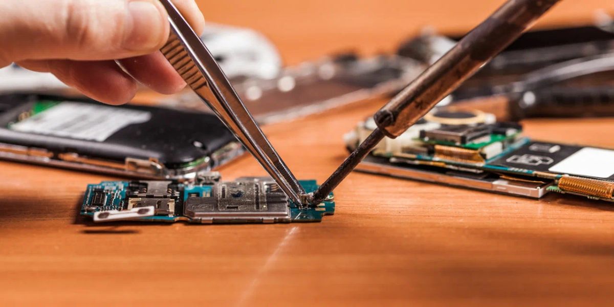 Close-up of a technician's hands repairing a small circuit board with tweezers and a soldering iron on a wooden desk