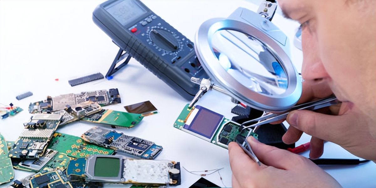Technician using a large magnifying glass and tweezers to inspect a small circuit board, with other boards and a multimeter nearby