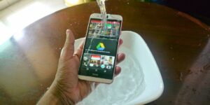 A hand holds an HTC smartphone while water is poured over it, with a white basin of water underneath
