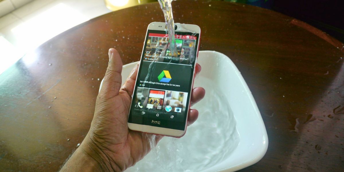 A hand holds an HTC smartphone while water is poured over it, with a white basin of water underneath