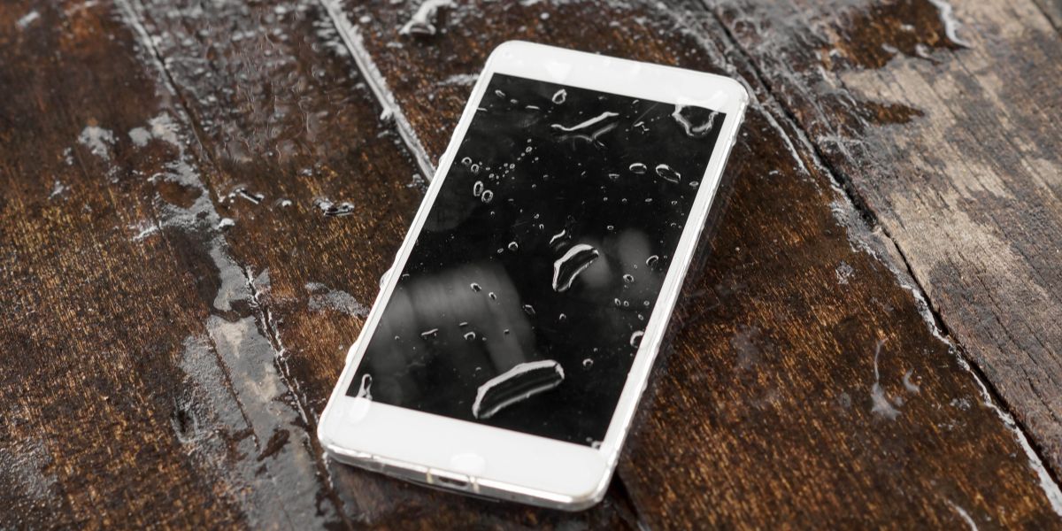 Wet white smartphone lying face-up on a dark, wet wooden table, showing droplets on the screen