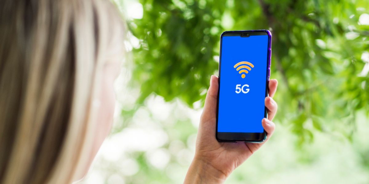 Woman outdoors holding a smartphone displaying an orange Wi-Fi icon and "5G" on a blue screen