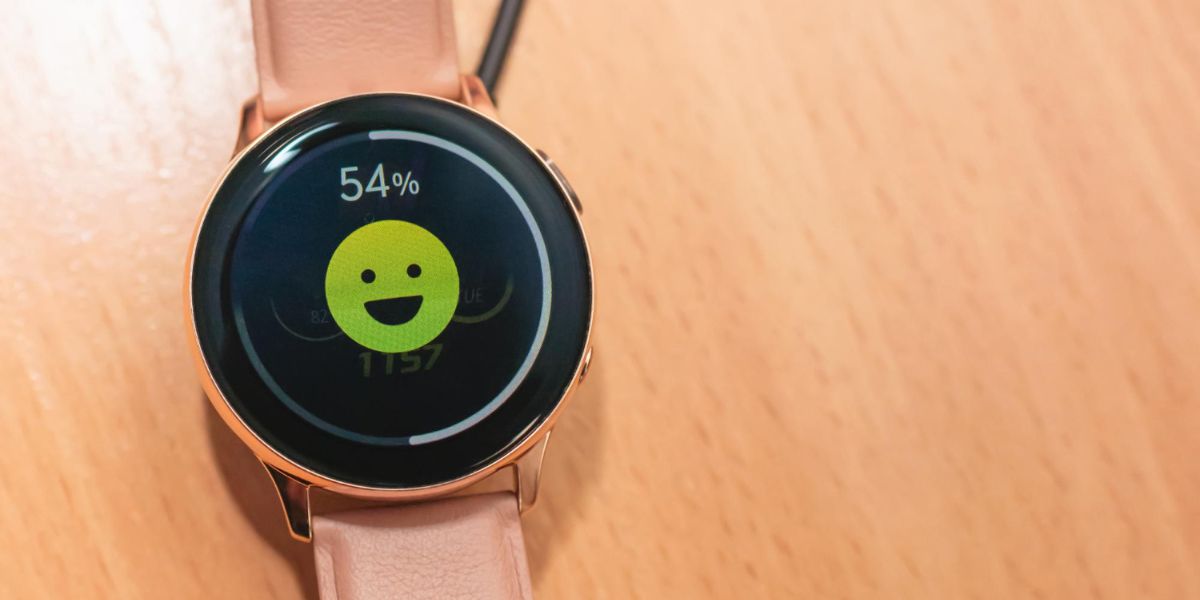 A close-up of a pink smartwatch displaying a 54% charge level and a large, smiling green face