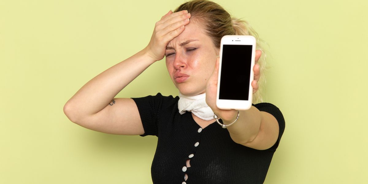 A frustrated young woman holds her hand to her forehead while showing a white smartphone with a blank black screen
