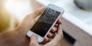 Close-up of a person's hands holding a white smartphone with a severely cracked and shattered glass screen outdoors