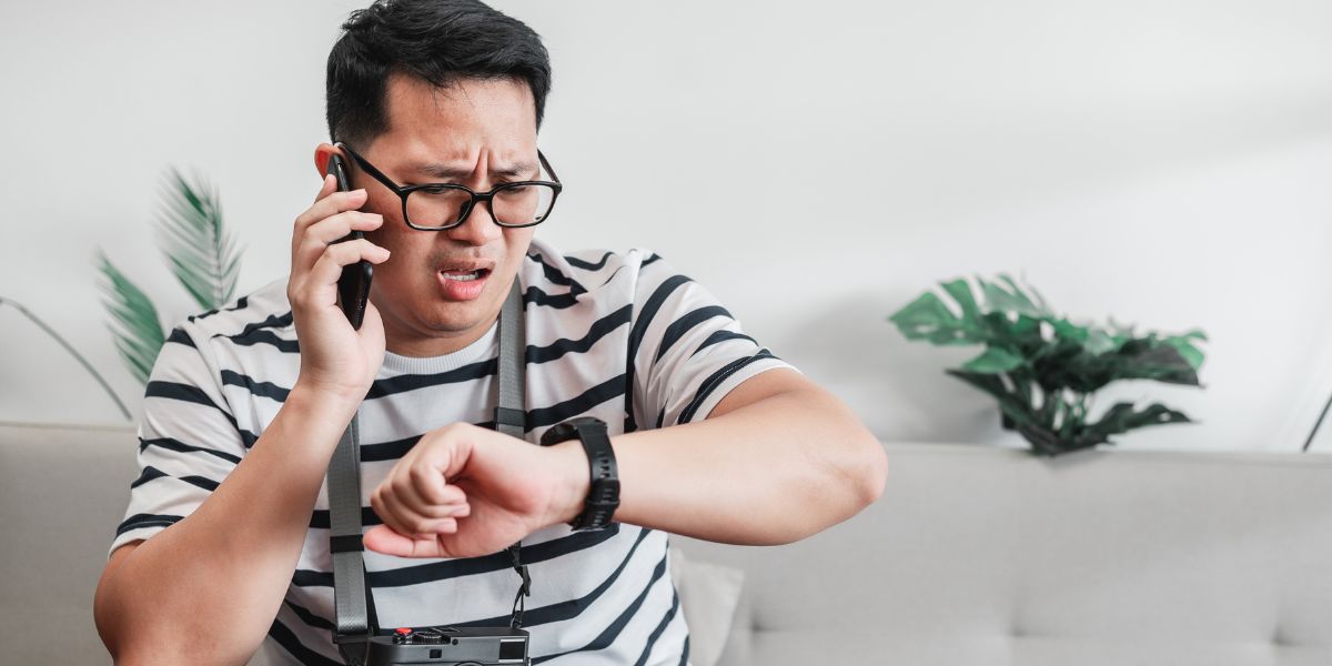 A man wearing glasses looks frustrated while checking his black smartwatch and talking on a smartphone indoors