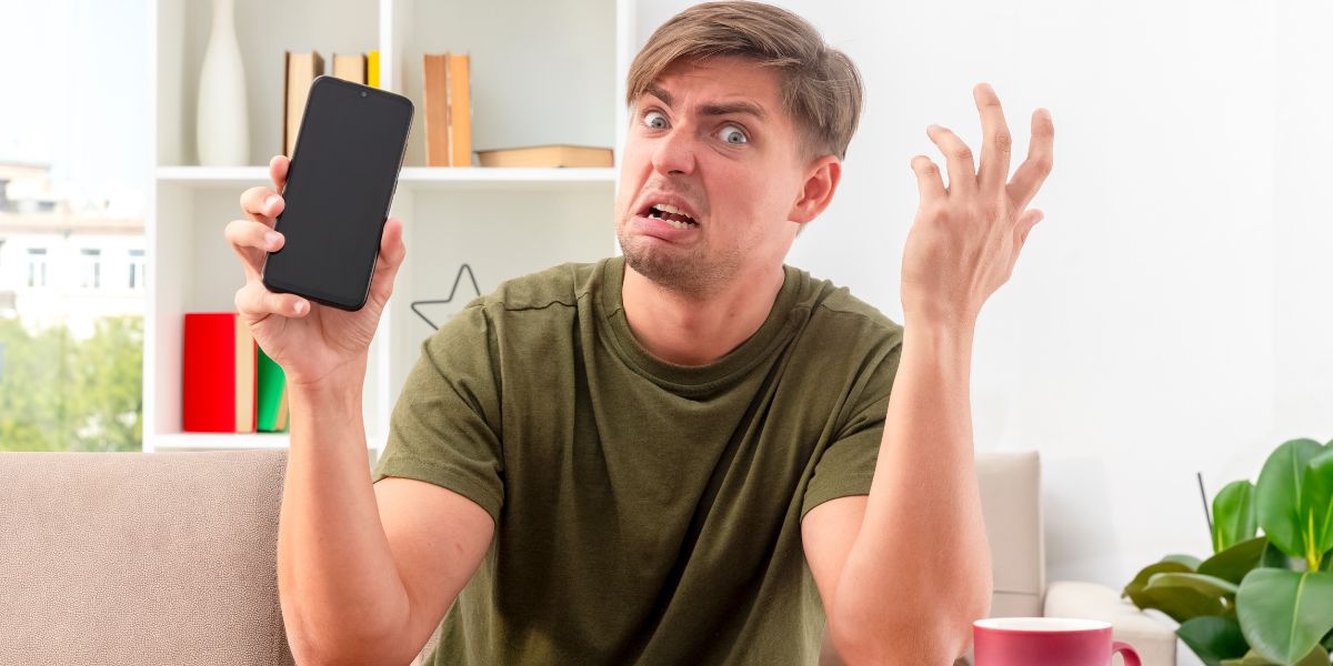 Frustrated man holding a black smartphone with a blank screen, gesturing with his hand in an annoyed expression